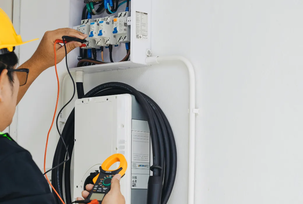 An electrician testing a charge point circuit at an open consumer unit using a clamp meter and test leads, with the wall-mounted charger below